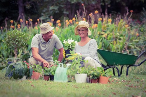 The image depicts an outdoor garden scene featuring two mature individuals, a man and a woman, seated in a well-maintained backyard with vibrant flower beds and lush greenery. The woman holds a small potted plant with white blossoms, smiling at the man, who appears engaged in conversation. Surrounding them are various garden tools and pots arranged on the grass, indicating ongoing gardening activities. In the background, a dense border of purple and orange flowers, along with green shrubs, creates a colourful backdrop. A green wheelbarrow is positioned behind the woman, suggesting recent planting or garden maintenance. The scene is illuminated by natural daylight under partly cloudy weather, contributing to a lively and inviting outdoor environment. The setting showcases typical features of a landscaped yard suitable for garden care and outdoor leisure, aligning with professional gardening and landscaping services offered by companies like Landscapers Wood Green, especially in the postcode area of Wood Green, London, N22.