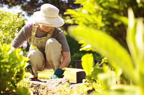 A gardener in a backyard garden at a sunny day, crouching down to tend to a flowerbed with a small gardening trowel. The garden features a well-maintained lawn with dense, green grass, bordered by neatly trimmed hedges and a variety of leafy shrubs. In the background, there are trees providing shade, and the garden includes a paved pathway made of stone slabs. The gardener is wearing a wide-brimmed hat, a patterned apron, and gardening gloves, emphasizing outdoor maintenance and landscaping activities typical of a residential garden in the Wood Green area near London. The scene highlights outdoor gardening work, with natural sunlight illuminating the space, supporting professional gardening and landscaping services by Landscapers Wood Green.