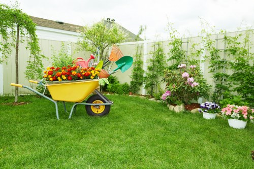 A well-maintained garden with a lush, green lawn in the foreground, bordered by flower beds and potted plants featuring colourful flowers such as pink, purple, and yellow blooms. A small decorative pond or container garden is visible among the foliage, while the background showcases a white privacy fence covered with climbing plants and vines. There is a yellow wheelbarrow filled with garden tools and supplies, including a watering can, garden rake, and small shovel, positioned on the grass. The garden appears to be in a residential outdoor space, likely in the Wood Green area, with a neat and organized layout suitable for gardening and outdoor maintenance activities. The environment is bright, suggesting a sunny day with diffused natural light. Guided by the principles of professional gardening services by Landscapers Wood Green, this garden demonstrates balanced plant placement and tidy surfaces, reflecting active care and attention to outdoor landscaping details.