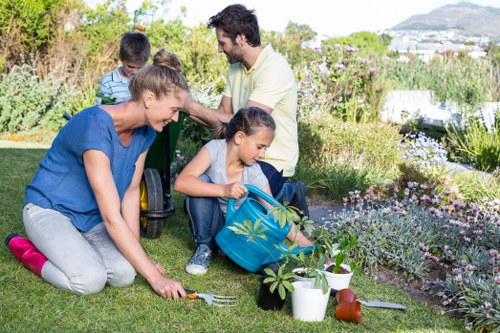 A family of four are engaged in gardening activities in a well-maintained backyard garden with lush green grass and a variety of flowering plants. The adult woman, dressed in a blue top and grey trousers, is kneeling on the grass, using a garden fork to tend to a small plant. A young girl, wearing a blue denim jacket, is sitting on the ground holding a small potted plant, with gardening tools and white pots nearby, indicating planting tasks. An adult man, standing behind them, is wearing a light yellow shirt and appears to be overseeing the activity, while a young boy in a striped shirt stands beside him, both observing the garden. The garden exhibits neatly defined flower beds with vibrant blooming flowers and foliage, bordered by a lawn area. In the background, there are mature hedges, shrubs, and possibly a patio or outdoor seating area, suggesting a spacious, landscaped outdoor environment typical of residential gardens in the Wood Green area. The weather appears clear and sunny, supporting outdoor garden work. This scene illustrates gardening services provided by Landscapers Wood Green, emphasizing family involvement and garden care in a picturesque outdoor setting, with an emphasis on healthy plant growth and landscape maintenance.