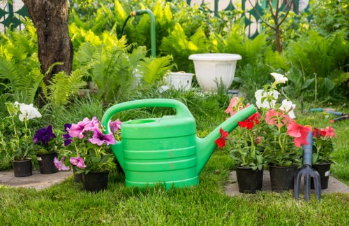 A garden scene with a well-maintained grassy lawn in the foreground, featuring a large green plastic watering can placed next to potted flowering plants in shades of pink, purple, and white. Behind these, there are various green shrubs, ferns, and small trees, creating a lush natural backdrop. To the right, a black hand fork is leaning against the plants, and in the background, a white garden planter with decorative patterns sits amidst the greenery. The scene appears to be part of a landscaped outdoor space, possibly within a residential garden near Wood Green, with natural daylight and mild weather conditions enhancing the vibrant colors of the plants and flowers. This setting aligns with outdoor gardening and landscaping services provided by companies like Landscapers Wood Green, emphasizing a well-kept and flourishing garden environment.