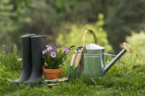 The image depicts a garden scene in a well-maintained outdoor space, likely in the Wood Green area, featuring a small flower bed with purple and white blooms in a terracotta pot, situated on lush green grass. Next to the flower bed are a pair of black rubber wellington boots placed upright, indicating garden ready footwear. Nearby, a set of gardening tools including a trowel, hand fork, and pruning shears are arranged on the grass. A traditional metal watering can with a curved spout and a brass nozzle sits beside the tools, suggesting ongoing or future plant care. The background shows a natural environment with trees and shrubs, creating a shaded, tranquil atmosphere typical of residential gardens in the region. The scene collectively highlights outdoor planting, watering, and general garden maintenance activities, aligning with expert gardening and landscaping services provided by Landscapers Wood Green. The lighting suggests a bright, clear day, contributing to the vibrant, inviting garden aesthetic in a typical suburban outdoor setting.