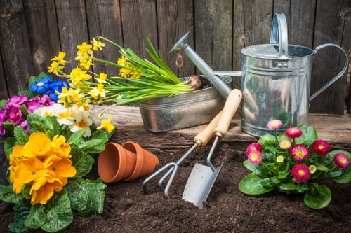 A newly prepared garden bed in a backyard outdoor space with dark, moist soil, surrounded by various garden tools and decorative plants. In the foreground, there are bright yellow and pink flowering plants with green leaves, adding vibrant colour to the scene. To the left, a cluster of potted plants and flower bulbs await planting, including an orange terracotta pot tipped over. Behind the planting area, a weathered wooden fence provides a natural backdrop. Several gardening tools, such as a small hand rake, trowel, and sprayer, rest on the soil surface, indicating active gardening work. The garden area is well-maintained, with a neat boundary between the soil and existing lawn, which is lush and green with a dense, healthy appearance. The scene suggests a temperate climate, typical of the Wood Green area, and reflects a focus on garden care and landscaping activities that a professional gardener or landscaper from Landscapers Wood Green might undertake to enhance outdoor spaces, including planting, soil preparation, and garden maintenance.