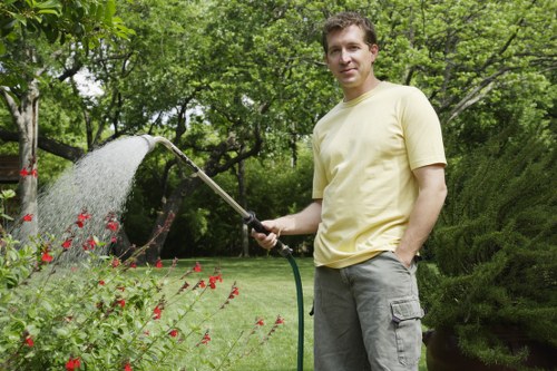 A man standing in a well-maintained garden lawn, holding a garden hose with water flowing onto the grass and flowering plants. The garden features a lush green lawn with dense, mature trees and bushes in the background, creating a natural and private outdoor space typical of front or back gardens in Wood Green. Nearby, there are flower beds with red and pink flowers, bordered by low soil edges, and a paved area visible in the background. The scene appears to be on a bright, mild day, with natural sunlight illuminating the garden and casting gentle shadows. The man is dressed casually in a pale yellow T-shirt and grey cargo shorts, engaged in outdoor maintenance, demonstrating professional gardening or lawn care activities that local landscapers like Landscapers Wood Green may provide. The overall environment emphasizes a vibrant, tidy, and inviting outdoor space suitable for gardening services focused on lawn care and garden maintenance in the local area.