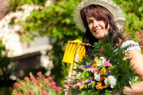 A woman standing outdoors in a garden, holding a large bouquet of colorful flowers including daisies, roses, and greenery. She is wearing a wide-brimmed straw hat and a white top, smiling at the camera. Behind her, there are lush green trees and vibrant pink flowering plants, with a well-maintained lawn visible in the foreground. The garden setting shows a clear, sunny day, suitable for outdoor gardening and landscaping activities. This scene illustrates the beauty of professionally maintained flower beds and outdoor spaces, aligning with services offered by companies like Landscapers Wood Green in the local area of Wood Green, blending natural garden elements with professional landscaping care.