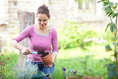 A woman in a pink and white striped long-sleeve shirt is tending to plants in a garden, holding a small terracotta pot with soil in her hands. She is working in a landscaped outdoor space that features a lush green lawn, with various flowering plants and shrubs in the foreground and background. The garden includes a paved patio area, possibly with paving stones or concrete, and a backdrop of trees and a wooden fence. The lighting indicates a natural, sunny day, contributing to a vibrant and well-maintained outdoor environment that reflects typical gardening activities supported by local landscapers in Wood Green. The scene emphasizes typical garden planting, soil preparation, and nurturing plants, aligning with gardening services offered in the area.