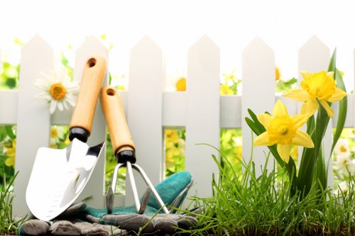 This image depicts a small garden space with a white picket fence in the background, set in a residential area near Wood Green, London. In the foreground, there is a patch of vibrant green grass and soil, with a few yellow daffodil flowers growing on the right side, adding bright natural colour to the scene. To the left, gardening tools are visible, including a trowel and a hand fork with wooden handles, resting on the ground. The garden appears to be well-maintained and is likely part of a landscaped outdoor area in a residential front or back garden. The lighting suggests a sunny day with clear weather, illuminating the lush greenery and blooming flowers. This scene reflects typical outdoor gardening activities and outdoor maintenance, which could be performed by professional landscapers such as Landscapers Wood Green, providing lawn care, planting, and garden upkeep services for local homes in the N22 postcode area.