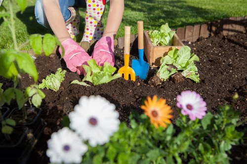 A close-up view of a gardener, wearing pink gardening gloves, planting young lettuce seedlings into dark, freshly turned soil within a flower bed in a backyard garden. Surrounding the planting area are colorful flowers, including white, orange, and purple blooms, positioned at the front of the image. In the background, a neatly maintained grassy lawn and a brick-edged border are visible, with small garden tools such as a yellow and blue trowel and a wooden plant label inserted into the soil. The outdoor environment appears bright and sunny, with natural lighting highlighting the vibrant green foliage and soil textures, reflecting typical features of gardening and outdoor landscape maintenance services in Wood Green.