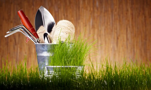 A close-up view of gardening tools and supplies arranged on a grassy lawn. The image features a metal bucket filled with various hand tools, including a small rake, garden trowel, pruning shears, and a spool of gardening twine, with some tools leaning against the bucket. The tools have wooden and metal handles, and the scene is set outdoors with a background of warm-toned wooden panels, indicating an outdoor garden or yard space. The grass in the foreground is lush, green, and dense, suggesting healthy lawn care, while the tools and setup imply ongoing gardening or landscaping activities typical of a residential garden in Wood Green, hinting at professional gardening services offered by Landscapers Wood Green. The natural lighting highlights the textures of the tools and the vibrant greenery, creating a professional and realistic depiction of garden maintenance preparations.