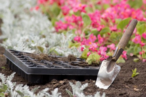The image shows a close-up view of a garden bed with dark, rich soil, partially covered by a black seed tray that contains some seedlings or soil preparations. In the foreground, a small gardening trowel with a wooden handle and metal blade is inserted into the soil, suggesting planting or soil aeration activity. Surrounding the workspace, there are lush green plants and small flowering plants with vibrant pink and white blossoms, indicating a well-maintained garden area. The background includes additional greenery and flowering plants, creating a colourful and inviting outdoor space typical of a landscaped garden in Wood Green. The scene is lit with natural daylight, reflecting an outdoor gardening environment, which aligns with professional gardening and landscaping services offered by Landscapers Wood Green, especially during spring or early summer seasons when planting activity is common.