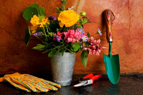 A grey concrete pot filled with a colorful arrangement of garden flowers, including yellow, pink, purple, and light blue blossoms, sits on a dark, textured outdoor surface. To the right of the pot, a small green trowel with a red handle is planted into the ground, while a pair of red-handled pruning shears and a small pair of secateurs rest nearby. On the left, a pair of yellow gardening gloves with orange and green stripes are laid flat. The background features a warm, reddish-brown textured wall, suggesting an outdoor garden or patio setting, with natural light illuminating the scene. The arrangement and tools indicate gardening activity typical of outdoor landscape maintenance in the local area served by Landscapers Wood Green, reflecting attention to floral decoration and garden upkeep, consistent with professional gardening services in the London Borough of Haringey.