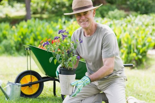 A cheerful elderly man wearing a straw hat, light grey t-shirt, and gardening gloves is kneeling on a well-maintained lawn in a landscaped outdoor space, holding a potted flowering plant with purple and red blossoms. Behind him, there is a black and yellow wheelbarrow partially filled with soil or garden debris, set on a lush green lawn with evenly cut grass. The background features a row of hedge bushes or shrubs, providing a natural boundary, and tall trees that offer dappled shade, indicating a sunny day with clear weather. The garden area appears tidy and landscaped, potentially part of a residential property serviced by local gardening specialists such as Landscapers Wood Green, who focus on lawn care, planting, and outdoor maintenance in this area. The scene emphasizes outdoor gardening and yard enhancement, with visible natural tones of green, brown, and vibrant plant colours, highlighting a well-kept garden environment suitable for approaching landscape or lawn care services.