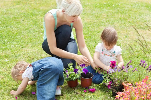 A woman with short blonde hair, dressed in a light blue top and dark jeans, kneels on a well-maintained lawn in a garden, engaging in planting activity with two young children. One child, wearing a white top and denim overalls, is bent over, closely inspecting or handling a flower in the grass. The other child, also in a white top, is seated nearby, holding purple and pink flowering plants. Several small terracotta pots and a black container filled with vibrant pink and purple flowers are arranged on the lawn. The garden features lush, green grass with a backdrop of hedges and a few trees, suggesting a neatly landscaped outdoor space typical of residential gardens in Wood Green. The scene is illuminated by natural daylight under partly cloudy skies, emphasizing the fresh, natural tones of the garden environment. Landscapers Wood Green specializes in gardening and landscaping services that enhance outdoor areas like this, with attention to plant selection, lawn care, and garden maintenance to create inviting and well-kept outdoor spaces.
