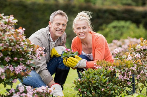 A man and a woman are kneeling in a well-maintained garden, surrounded by vibrant flowering shrubs with pink and white blooms. The garden features a neatly trimmed lawn in the foreground, with lush green bushes and flower beds extending into the background. The man is wearing a light grey shirt and gloves, holding a gardening tool, while the woman is dressed in an orange cardigan and yellow gardening gloves, both smiling at the camera. The scene is set outdoors in a bright, clear day with natural sunlight, and the garden is organized with defined borders and healthy, colorful plants, reflecting professional landscaping and gardening care typical of a residential garden in Wood Green, London, or similar areas. This setting showcases outdoor maintenance, planting, and garden enhancement services provided by companies like Landscapers Wood Green, emphasizing the importance of proper garden upkeep and safety in outdoor spaces.